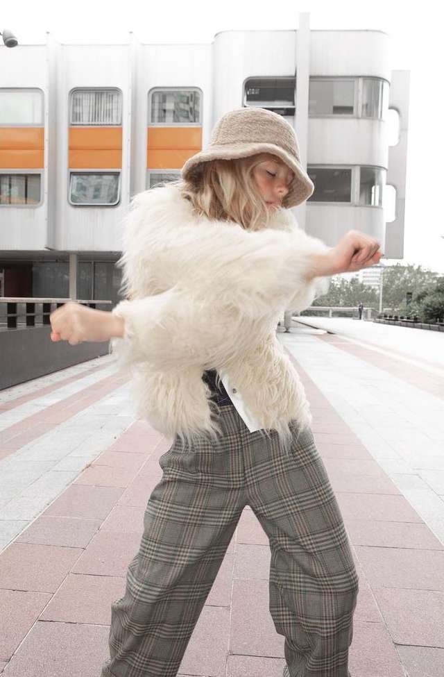 Adult female in furry white jacket and plaid pants dances in an urban setting.  She wears a knitted hat in front of a modern building.