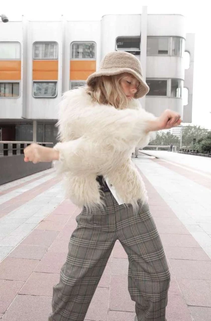 Adult female in furry white jacket and plaid pants dances in an urban setting.  She wears a knitted hat in front of a modern building.