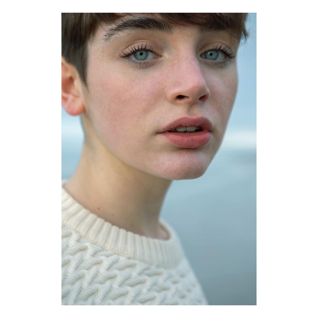 Close-up portrait of a young adult woman with short brown hair and blue eyes, wearing a white textured sweater, against a blurred backdrop.
