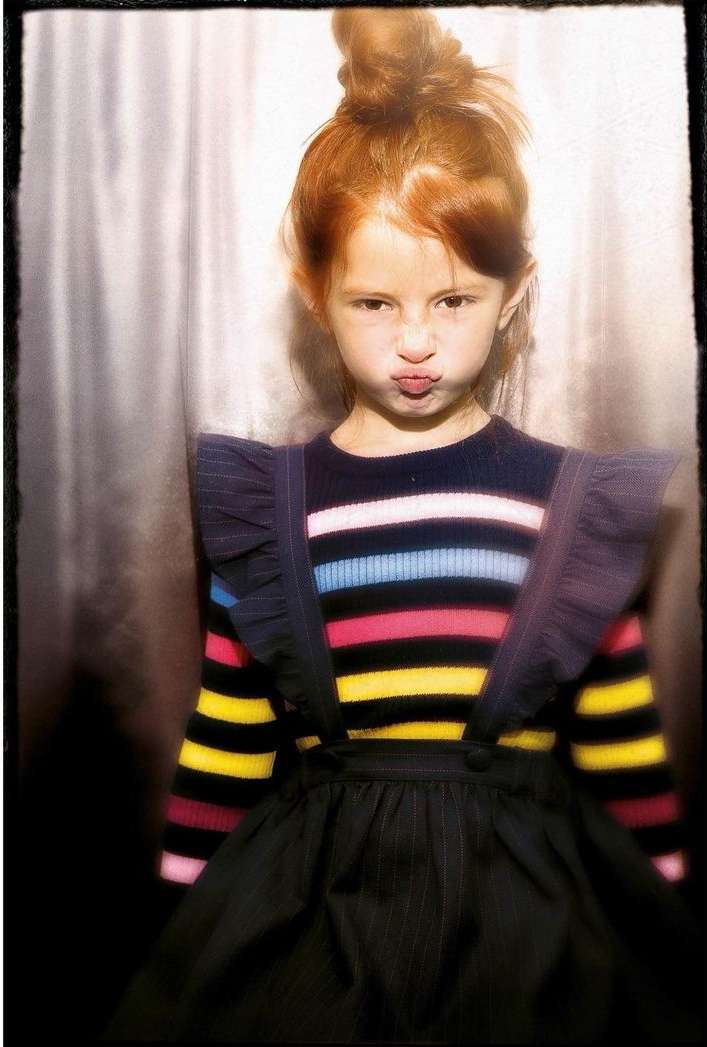 Portrait of a red-haired child with pouting lips wearing a striped top and black pinafore dress against a grey backdrop.