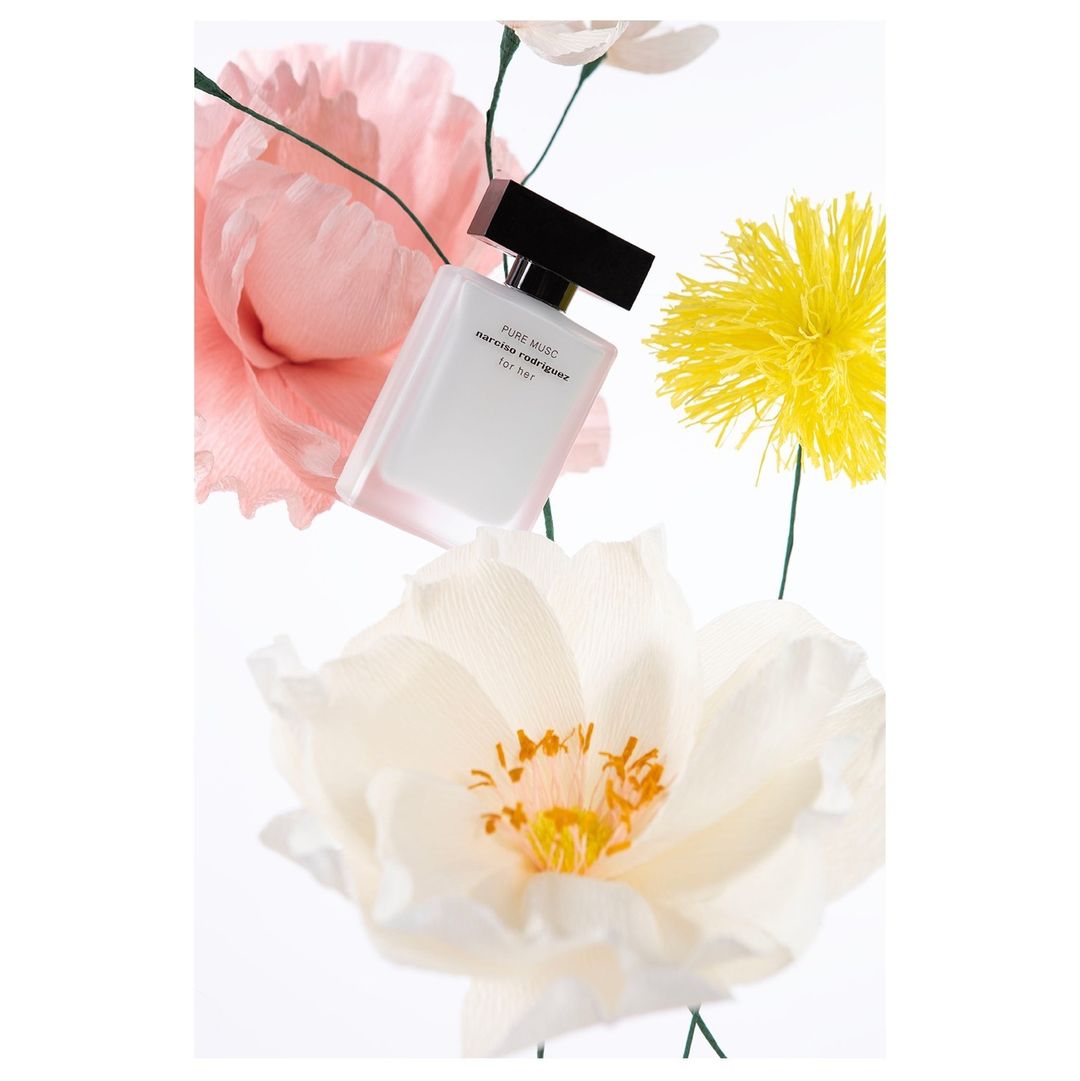 Still life photograph of a bottle of perfume surrounded by decorative paper flowers in white, pink, and yellow against a bright white background.
