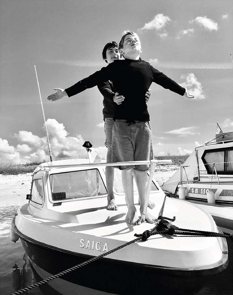 Two boys recreate the iconic Titanic pose on a small boat under a cloudy sky in a black and white, full shot, outdoor setting.