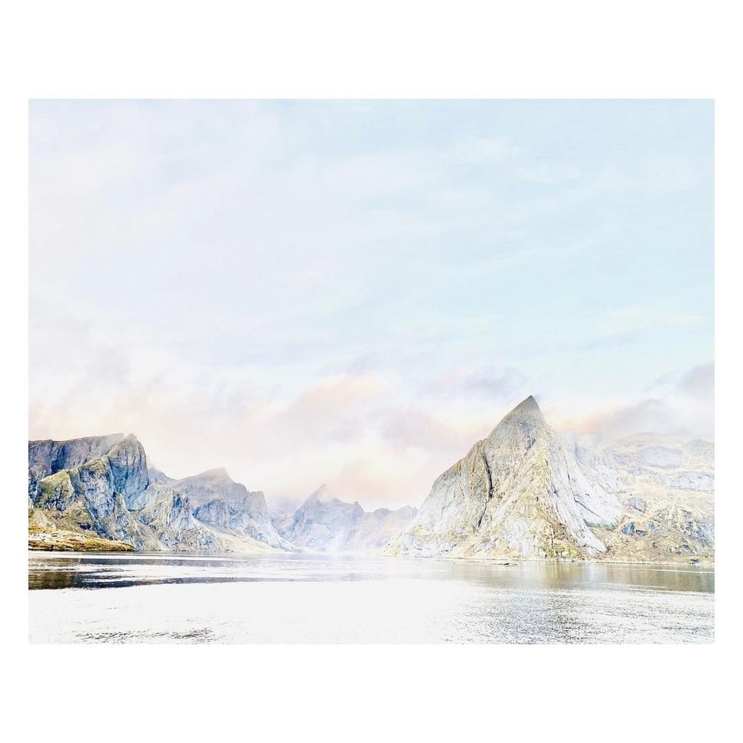 Landscape view of a reflective body of water surrounded by misty, rocky mountains under a light sky, showcasing a serene nature scene.