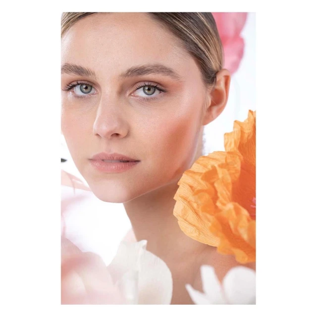 Close-up portrait of a woman with neutral makeup, framed by decorative paper flowers in pink, peach, and white against a clean white background.