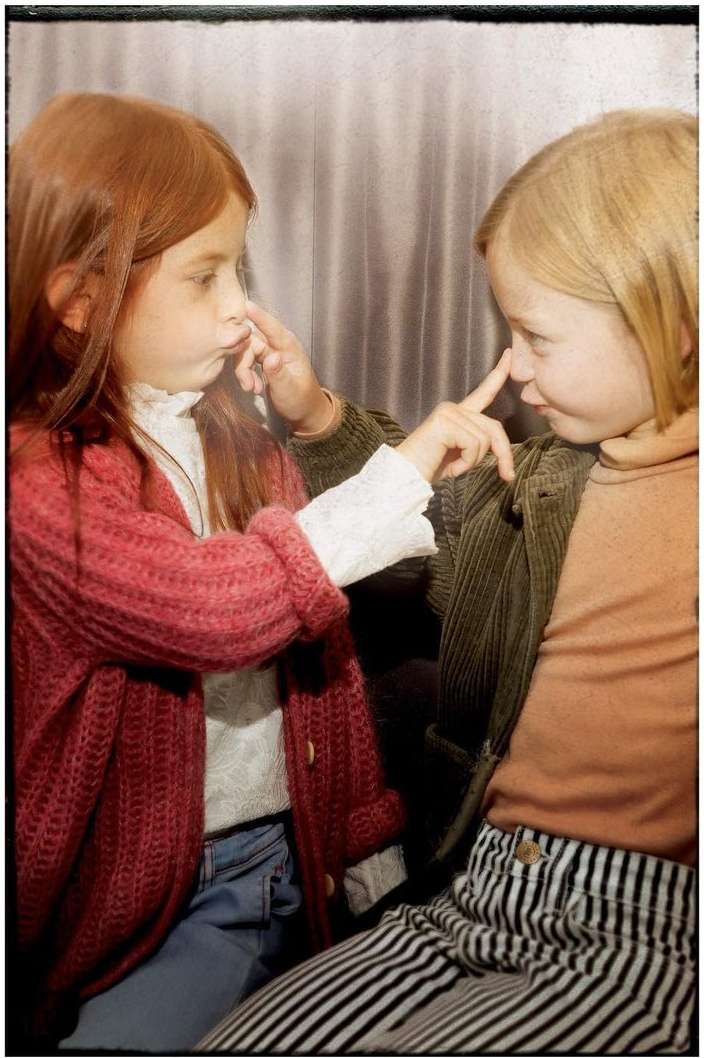 Two young girls with fair complexions face each other, playfully touching noses in an indoor setting with muted color grading.