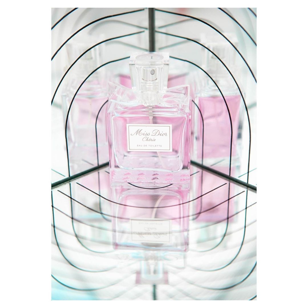 Close-up of a bottle of Miss Dior Cherie Eau de Toilette with a clear cap, set against a mirrored background that creates multiple reflections.