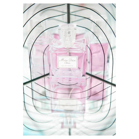 Close-up of a bottle of Miss Dior Cherie Eau de Toilette with a clear cap, set against a mirrored background that creates multiple reflections.