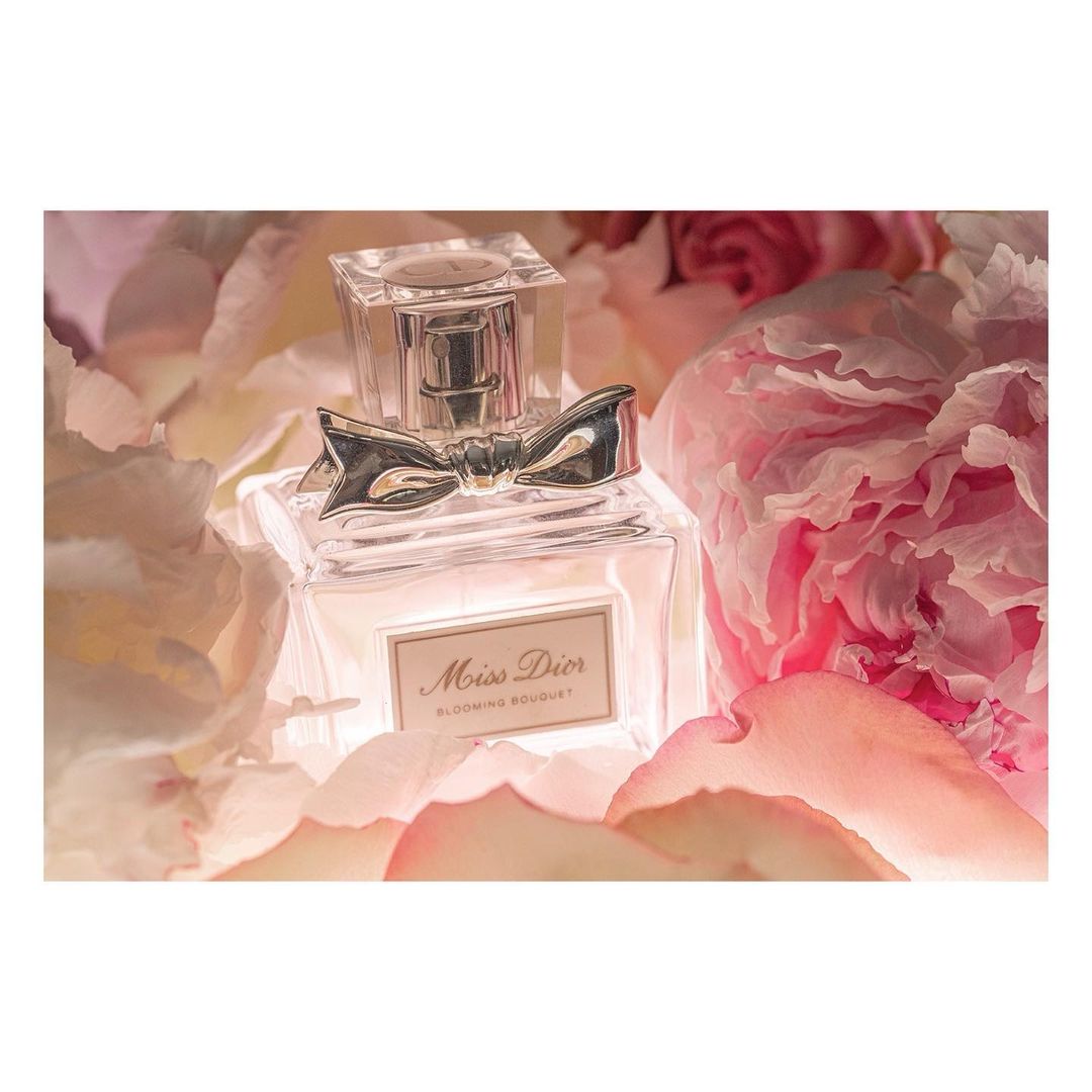 Close-up of a Miss Dior Blooming Bouquet perfume bottle surrounded by soft pink and white peony petals in a well-lit, elegant beauty shot.