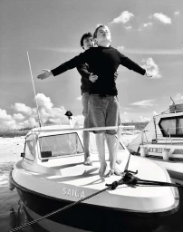 Black and white portrait of two boys standing on the bow of a small moored boat under an open sky.