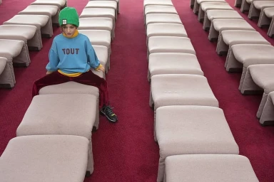 Child wearing green beanie and blue sweatshirt sitting among rows of padded stools on red carpet
