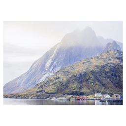 A towering mountain partially shrouded in mist rises behind a rocky coastal landscape and calm water by Sophie Le Gendre.