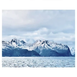 Snow-dusted mountain range rising directly behind a calm expanse of deep blue water under an overcast sky.