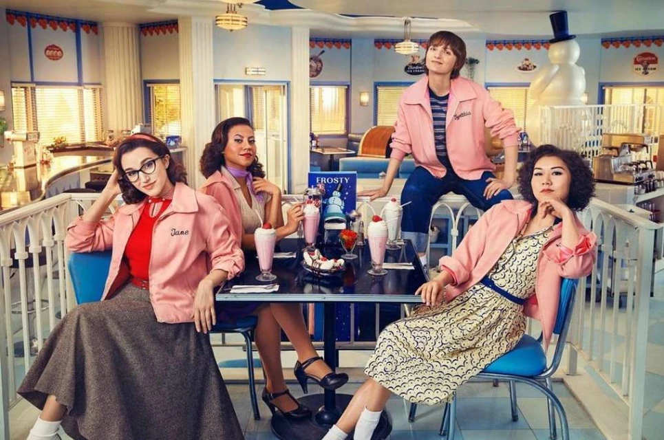 Four women in retro diner setting wearing pink jackets, sitting at a table with milkshakes and desserts, smiling and posing for camera.