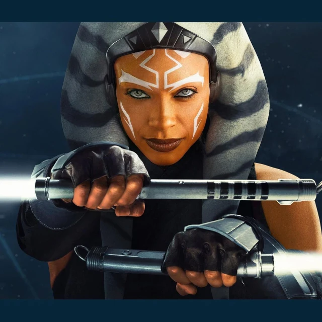 Close-up of Ahsoka Tano character holding dual lightsabers, with orange skin, white markings, and grey headwear on a blue background.