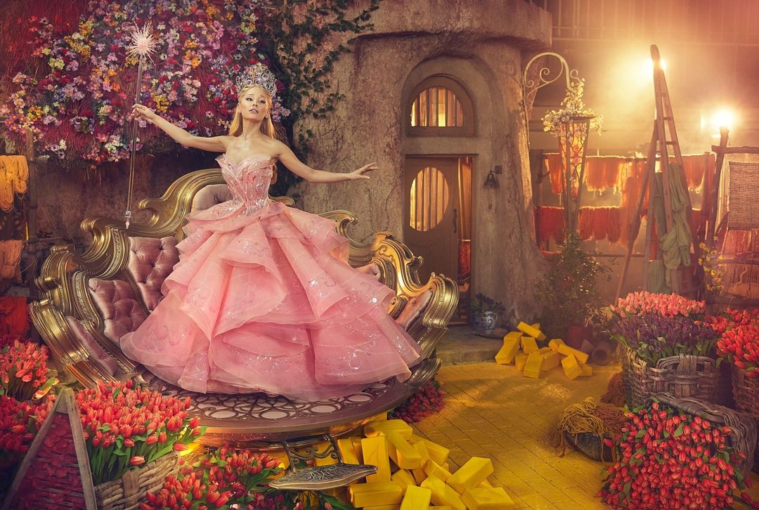 Woman in a pink gown and crown sits on an ornate gold bench surrounded by red tulips and gold bars in front of a stone building.