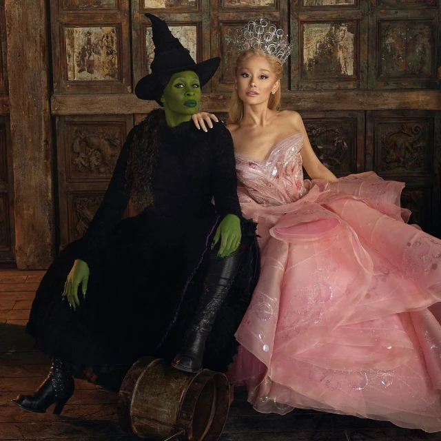 Two women posing together, one with green skin and a witch hat, the other in a pink gown and tiara, with vintage door panel background.