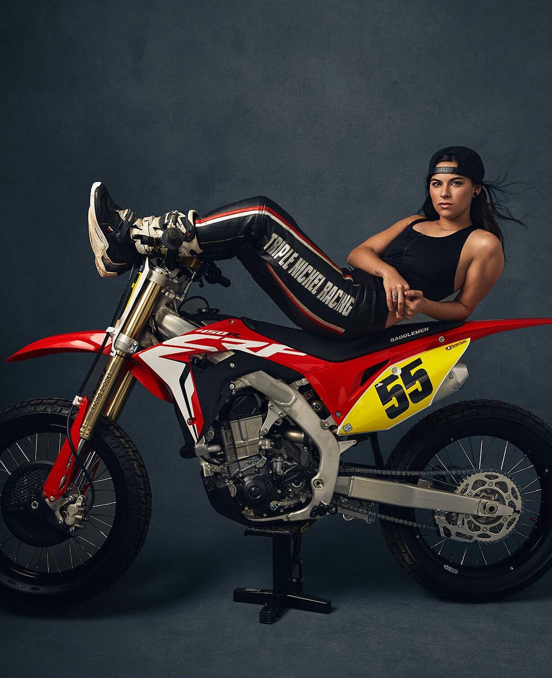 Adult woman poses on a red and white dirt bike indoors in a photo studio with a gray backdrop, wearing black tank top and leather pants.