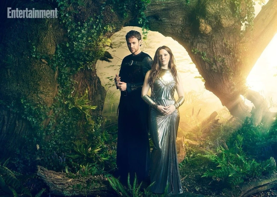 A man and woman pose in a forest. The man wears a black tunic, and the woman wears a silver dress and armbands in a magical setting.