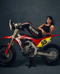 Woman in black tank top and riding pants lounging on a red dirt bike against a dark textured studio backdrop.