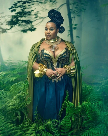Black woman in elaborate green cape and gold jewelry standing amidst lush ferns in a misty forest by Sophy Holland
