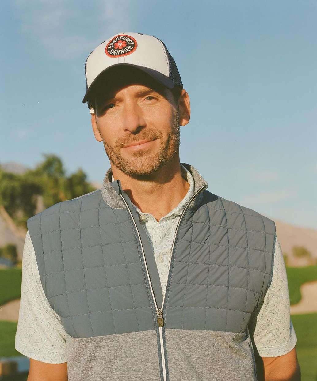 Portrait of an adult male wearing a baseball cap and a quilted vest outdoors on a sunny day with a blue sky in the background.