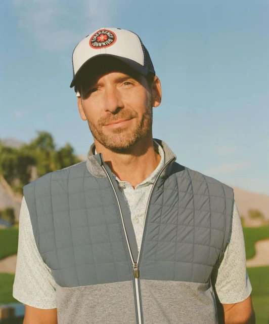 Portrait of an adult male wearing a baseball cap and a quilted vest outdoors on a sunny day with a blue sky in the background.
