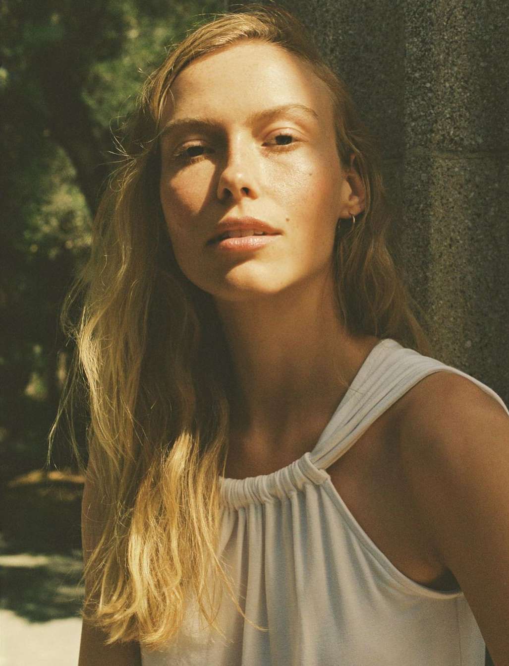 Close-up portrait of a blonde woman in a white top, standing outdoors with diffused natural light and blurred background foliage.