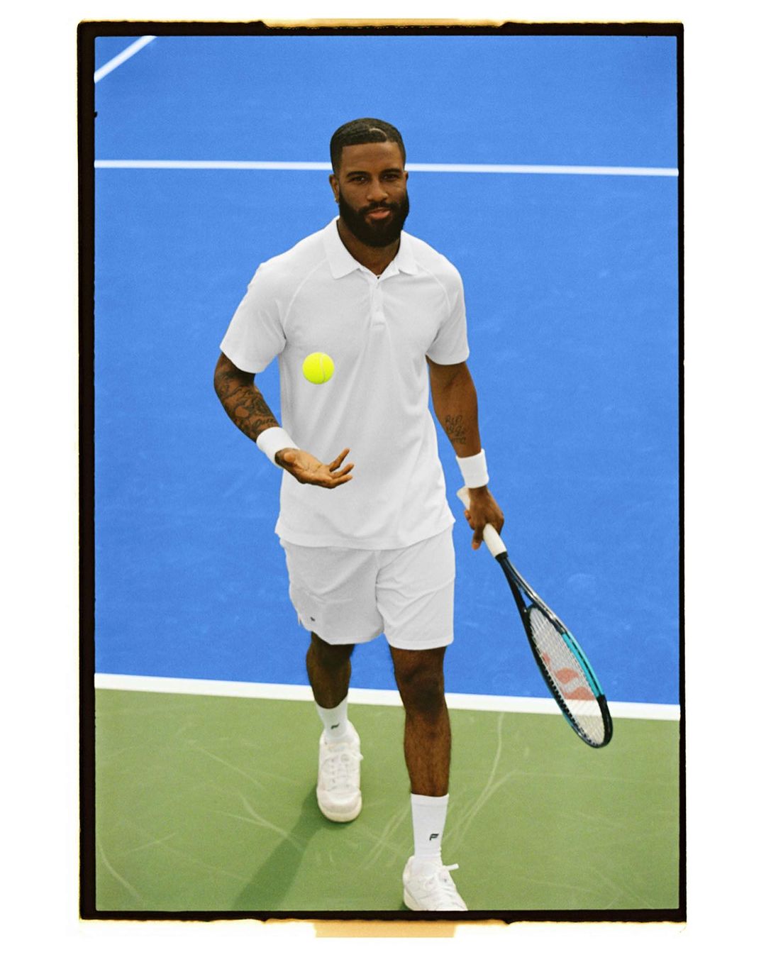 Athletic man in white tennis apparel on a blue and green court tossing a tennis ball, holding a racquet, and looking at the camera.
