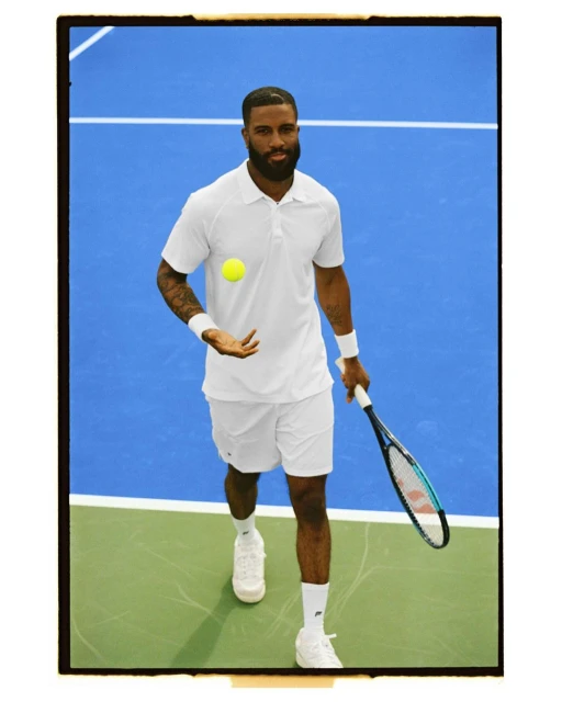 Athletic man in white tennis apparel on a blue and green court tossing a tennis ball, holding a racquet, and looking at the camera.
