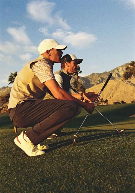 Two golfers in caps crouch on a green course with mountain views. They hold golf clubs, enjoying a clear, sunny day on the green.
