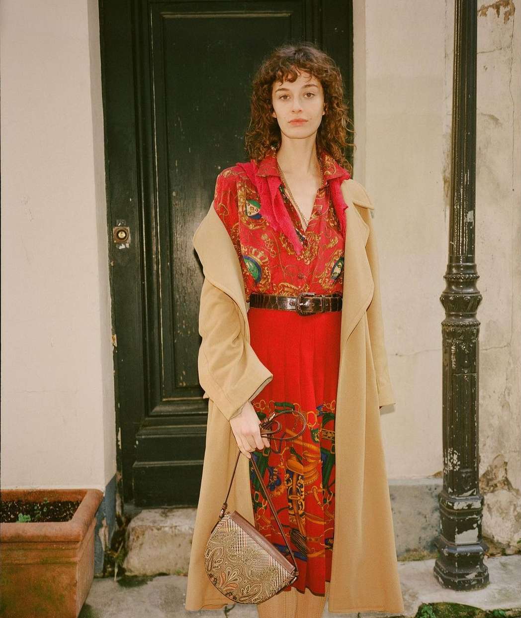 Woman with curly brown hair wearing a red patterned dress, camel coat, and holding a handbag in front of a dark door.