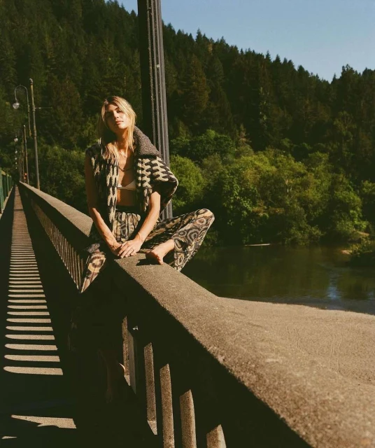 Woman in patterned vest and pants sits on a concrete bridge overlooking a tree-lined river in a sunny, outdoor fashion shot.