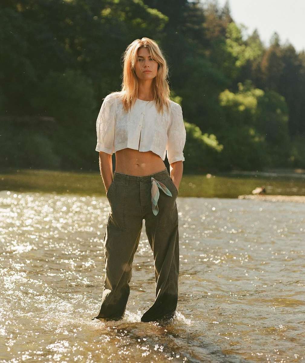 Blonde woman in a white top and olive pants stands in a flowing river with a forest backdrop under natural light, outdoors.