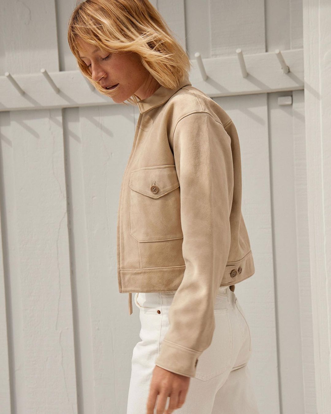 Woman poses in a short tan suede jacket and white jeans against a white wall, showcasing fashion and style in a neutral setting.