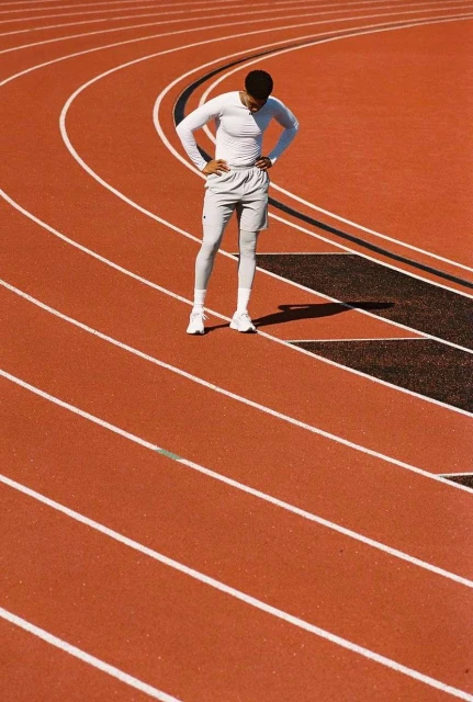 Athletic man pauses on a red running track, hands on hips, wearing a white long-sleeved shirt, athletic shorts, and white running shoes.