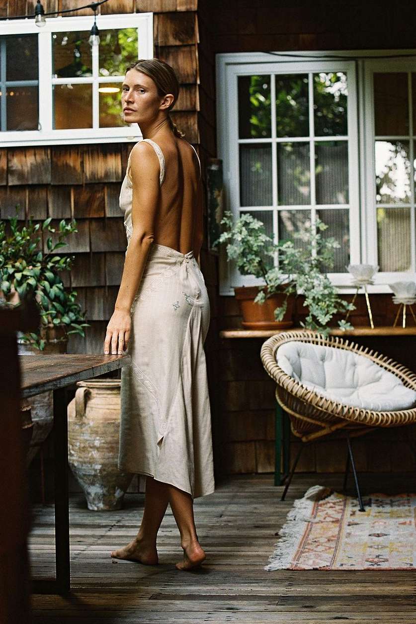 Woman in a sleeveless dress looks over her shoulder in a rustic outdoor setting with plants and a chair. Light wood textures.