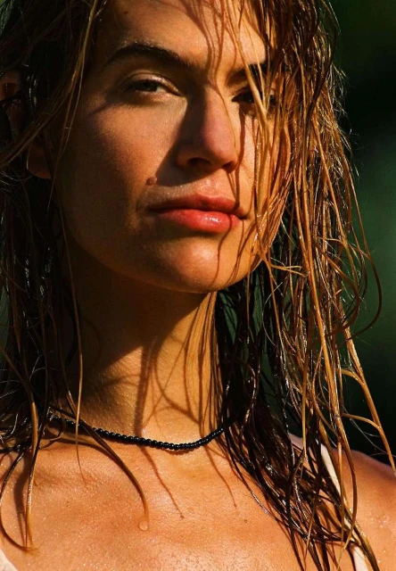 Close-up portrait of a woman with wet blonde hair and a black beaded necklace, bathed in sunlight against a blurred green background.