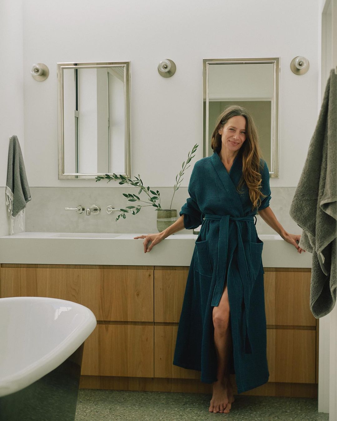A woman in a blue robe stands in a well-lit bathroom with a modern sink, mirror, and green-toned floor. Bathtub is partially visible.