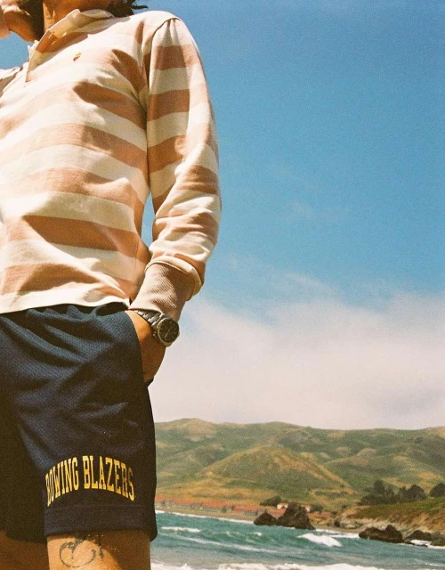 A person in a striped shirt and athletic shorts stands near a beach with distant mountains under a clear sky, showcasing casual style.