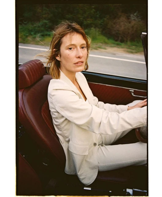 Woman in a cream-colored suit driving a vintage red convertible along an open road with a blurred natural background.