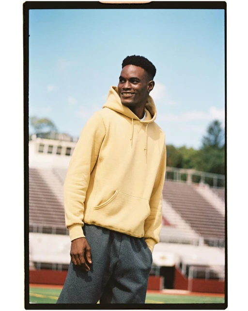 A young adult Black man in sportswear smiles outdoors. He wears a yellow hooded sweatshirt and grey sweatpants in a sports setting.
