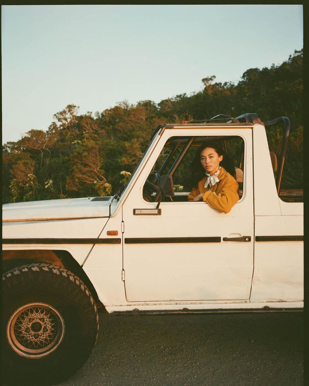 Young woman drives white SUV along a paved road with trees in background. She wears a yellow jacket and white neck scarf.