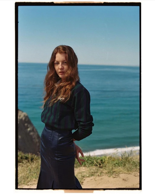 Young woman with auburn hair poses in front of the ocean, wearing a striped sweater and leather skirt on a sunny day.