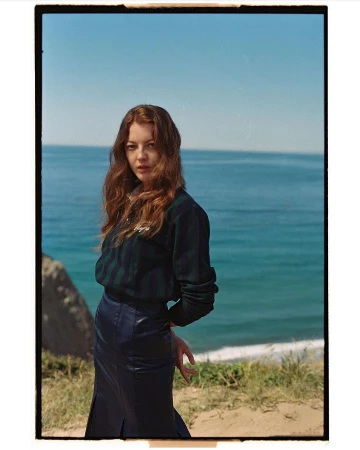 Young woman with auburn hair poses in front of the ocean, wearing a striped sweater and leather skirt on a sunny day.