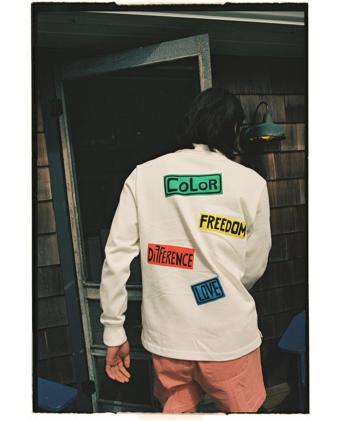 Back view of a person wearing a white long-sleeve shirt with colorful word patches and salmon shorts standing near a weathered door.