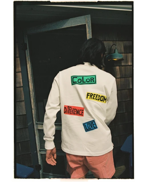 Back view of a person wearing a white long-sleeve shirt with colorful word patches and salmon shorts standing near a weathered door.