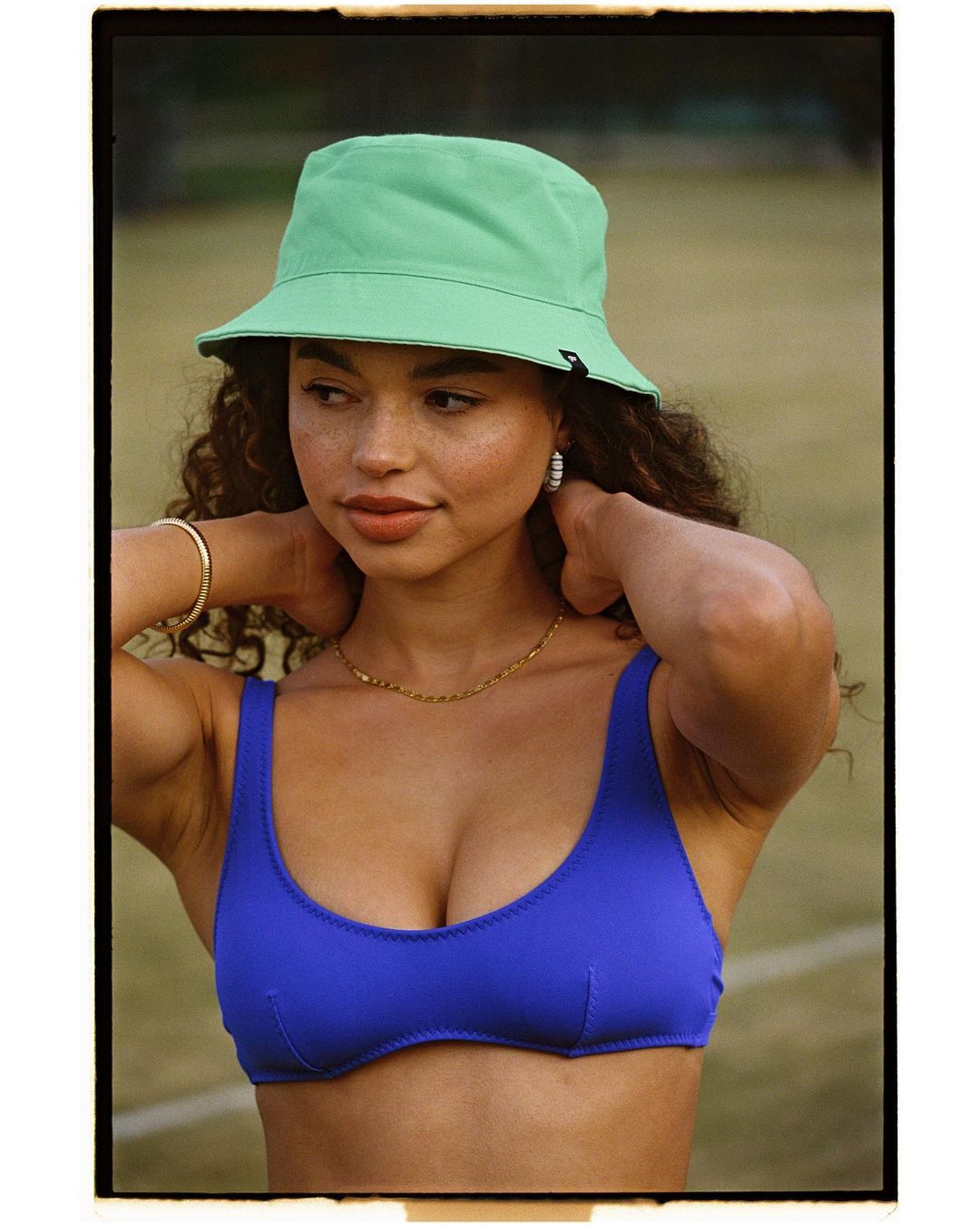 Adult female poses wearing a blue bathing suit top and light-green bucket hat in a medium shot with blurred background.