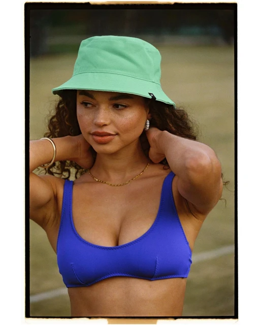 Adult female poses wearing a blue bathing suit top and light-green bucket hat in a medium shot with blurred background.