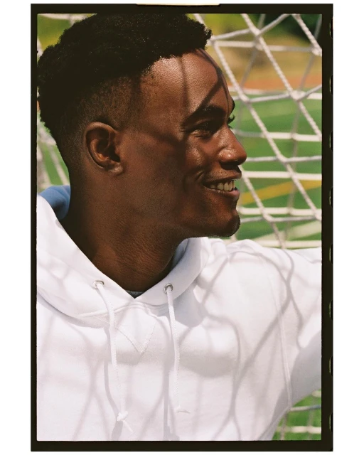 Profile portrait of smiling African-American man in white hooded sweatshirt, standing in front of a blurred soccer net outdoors.