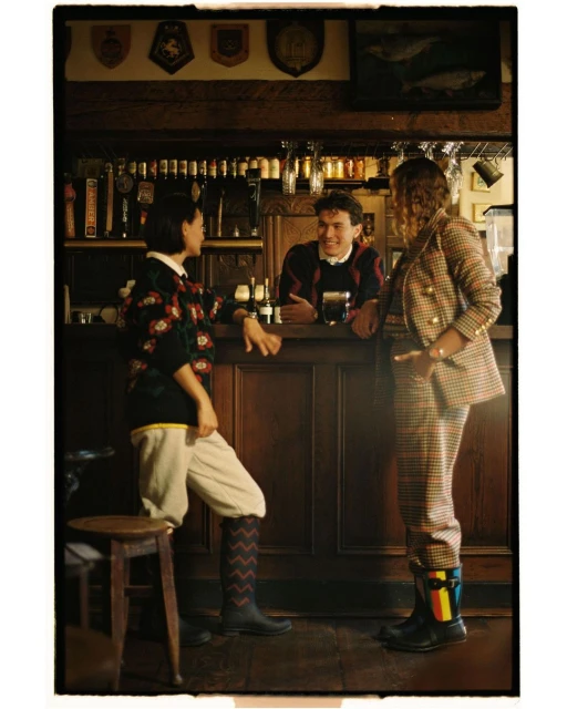 Three people in vintage-inspired clothing stand at a bar with dark wood, enjoying drinks in a photograph with warm, inviting lighting.
